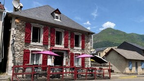 Un edificio de piedra con un letrero de restaurante, mesas al aire libre con sombrillas y una montaña al fondo.