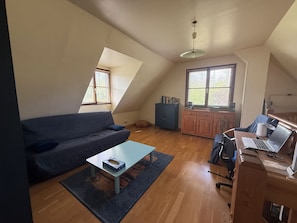 A loft-style room with a sloped ceiling, a sofa, a coffee table, a desk with a laptop, and a window with blinds. - Les Buissons (st pierre le vieux)