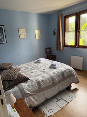 A bedroom with a bed, a chair, a window with curtains, and wall art. - Les Buissons (st pierre le vieux)