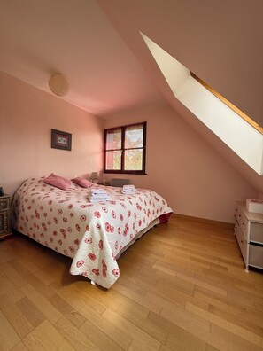 Ein Schlafzimmer mit schrägen Wänden, einem Bett mit floralem Bettbezug, einem Nachttisch und einem Fenster mit Jalousien.