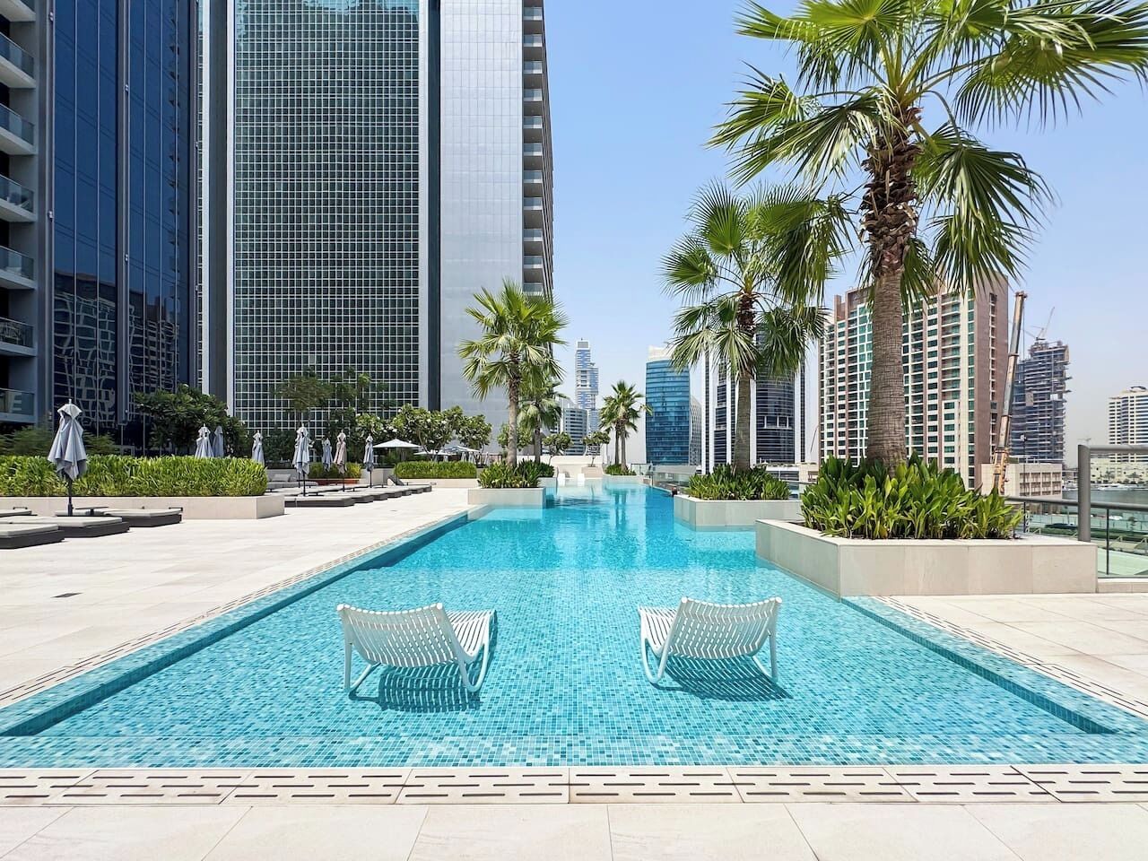 A swimming pool with lounge chairs, surrounded by palm trees and modern buildings.