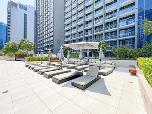 A rooftop lounge area with sunbeds and umbrellas, surrounded by modern buildings. - Chic 1BR Duplex in The Sterling | Business Bay (Dubai)