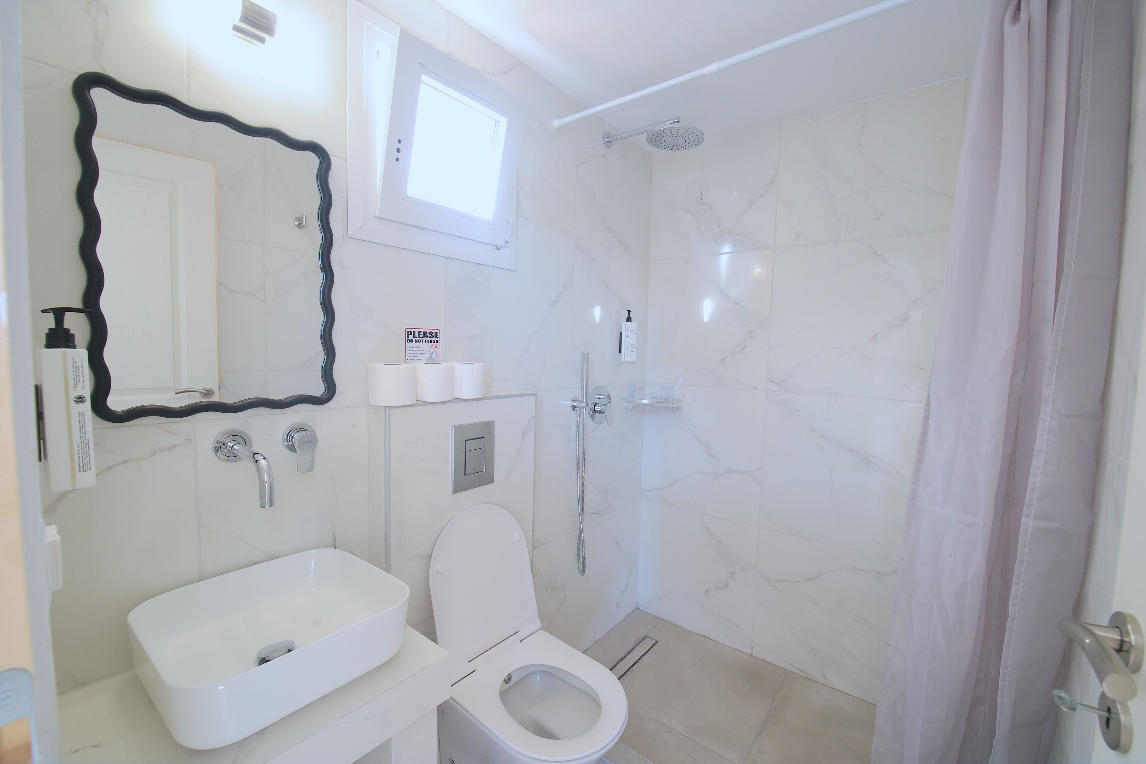 Un baño con lavamanos, inodoro y ducha blancos. Hay un espejo con marco negro y un cartel en la pared.