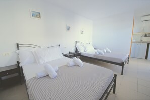 A hotel room with two beds, each with a pillow and a rolled towel. There is a small bedside table and a wall-mounted shelf. - Tinamar Selection (Santorini)