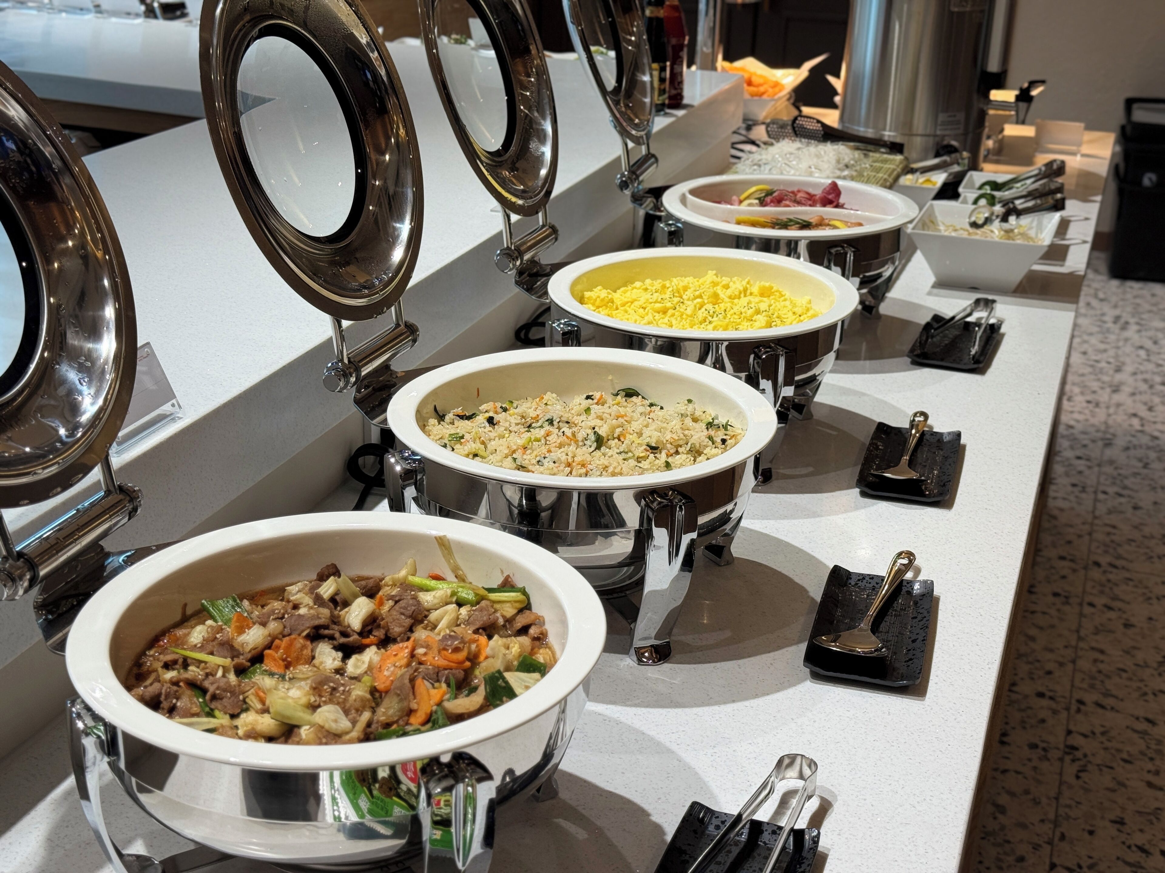 Un bufé con múltiples recipientes de comida al servicio, incluyendo platos de arroz y verduras salteadas.