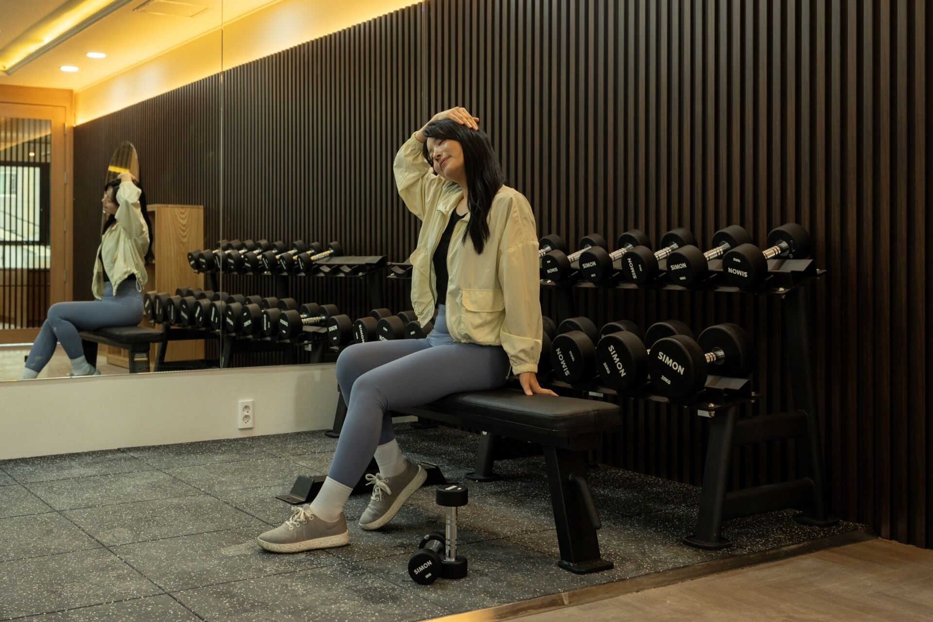 Eine Person sitzt auf einer Bank in einem Fitnessstudio mit Spiegel und Reihen von Kurzhanteln.