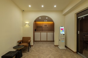 Un espace d’accueil d’hôtel comprenant un comptoir de réception, un coin salon avec une chaise et une table, et un kiosque en libre-service.