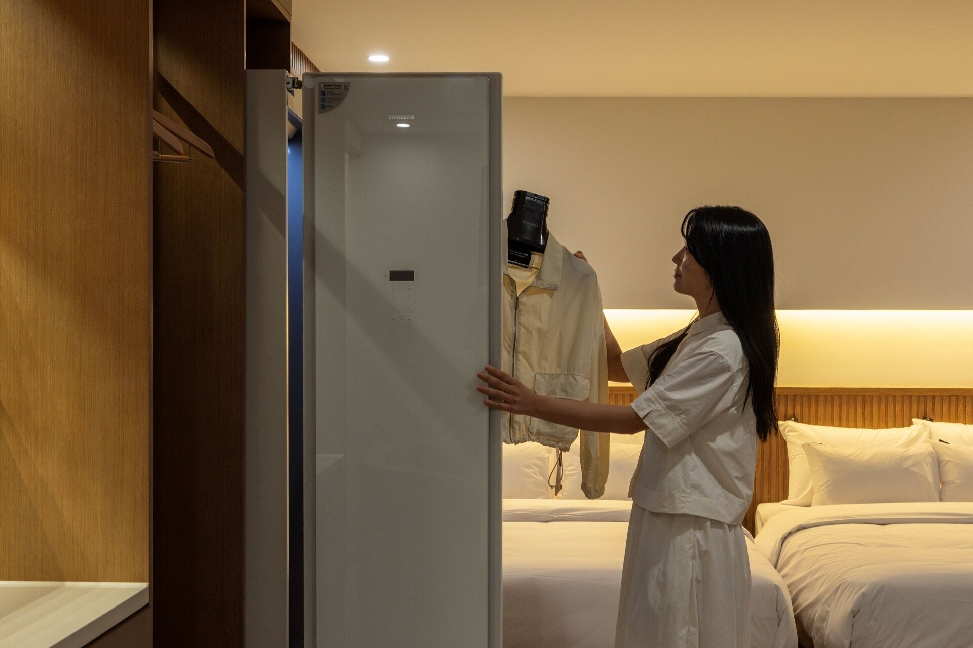 Una persona con camisa y falda blanca de pie junto a un refrigerador blanco en una habitación de hotel con cama y cabecera de madera.