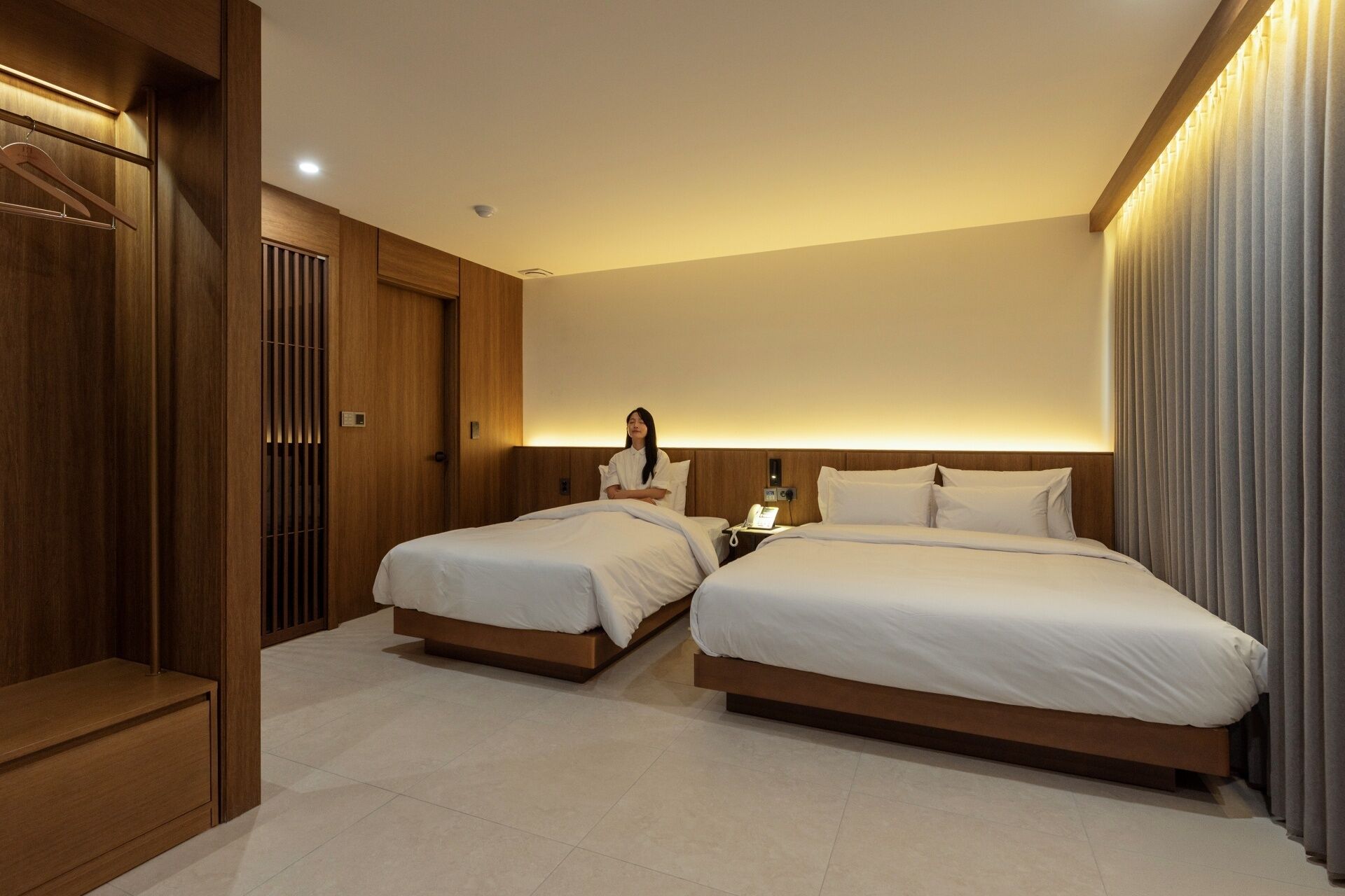 A hotel room with two beds, a wooden wardrobe, a bedside table with a lamp, and a wall-mounted phone.