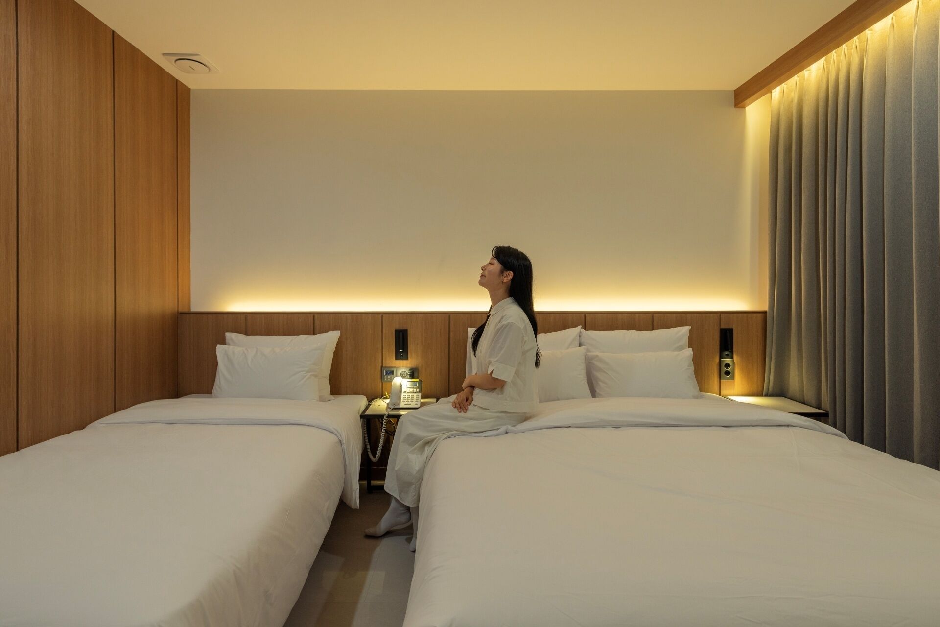 A hotel room with two beds, a bedside table with a phone, and a headboard with built-in lighting.