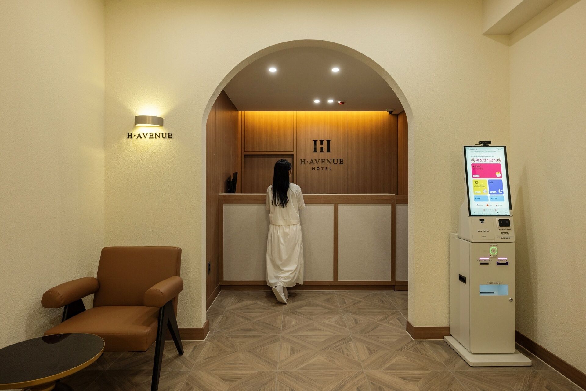 A hotel reception area with a woman approaching the desk, a seating area with a chair, and a self-service kiosk.