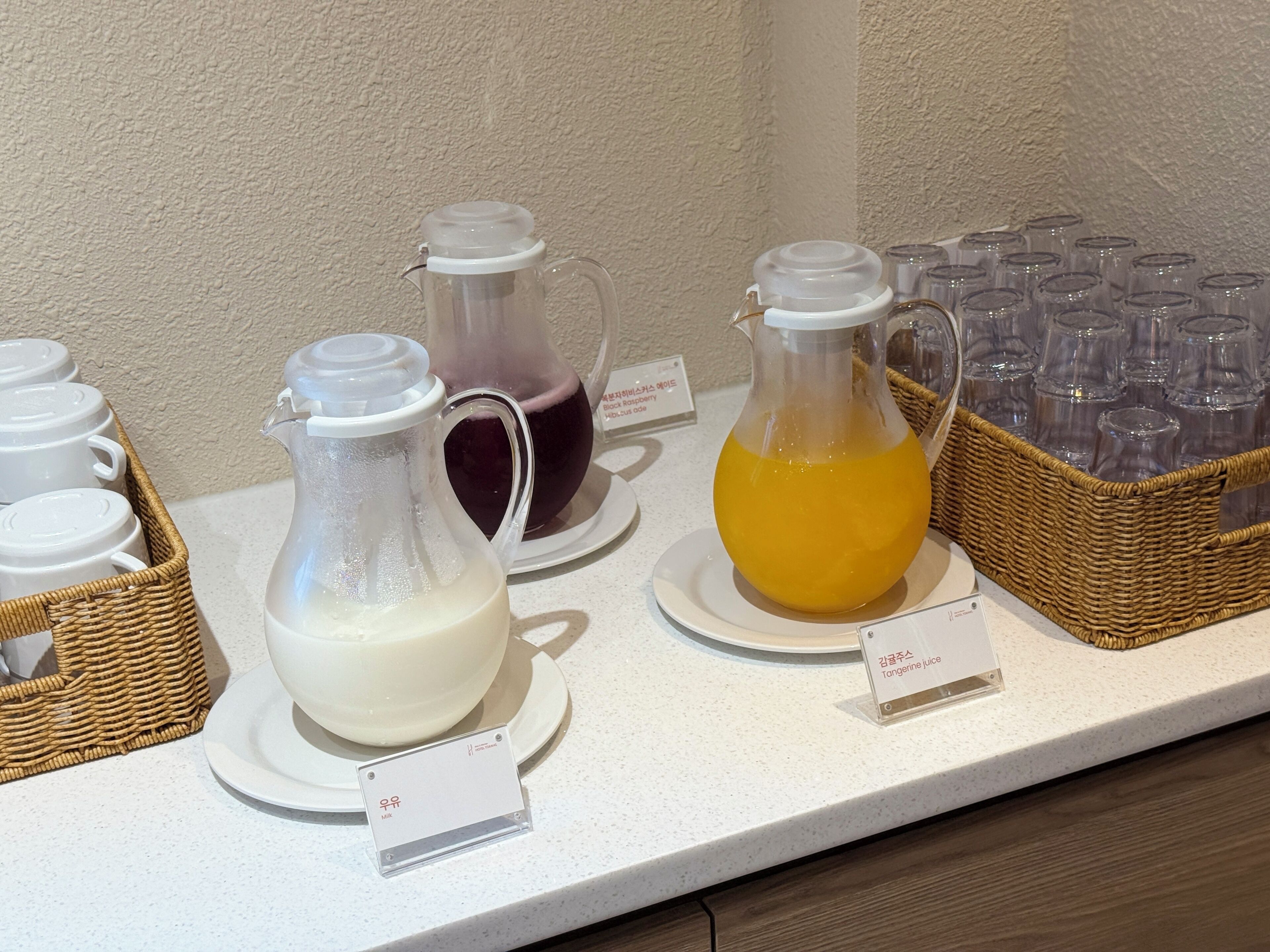 A beverage station with three pitchers of different drinks, a basket of glasses, and a sign indicating the type of juice.
