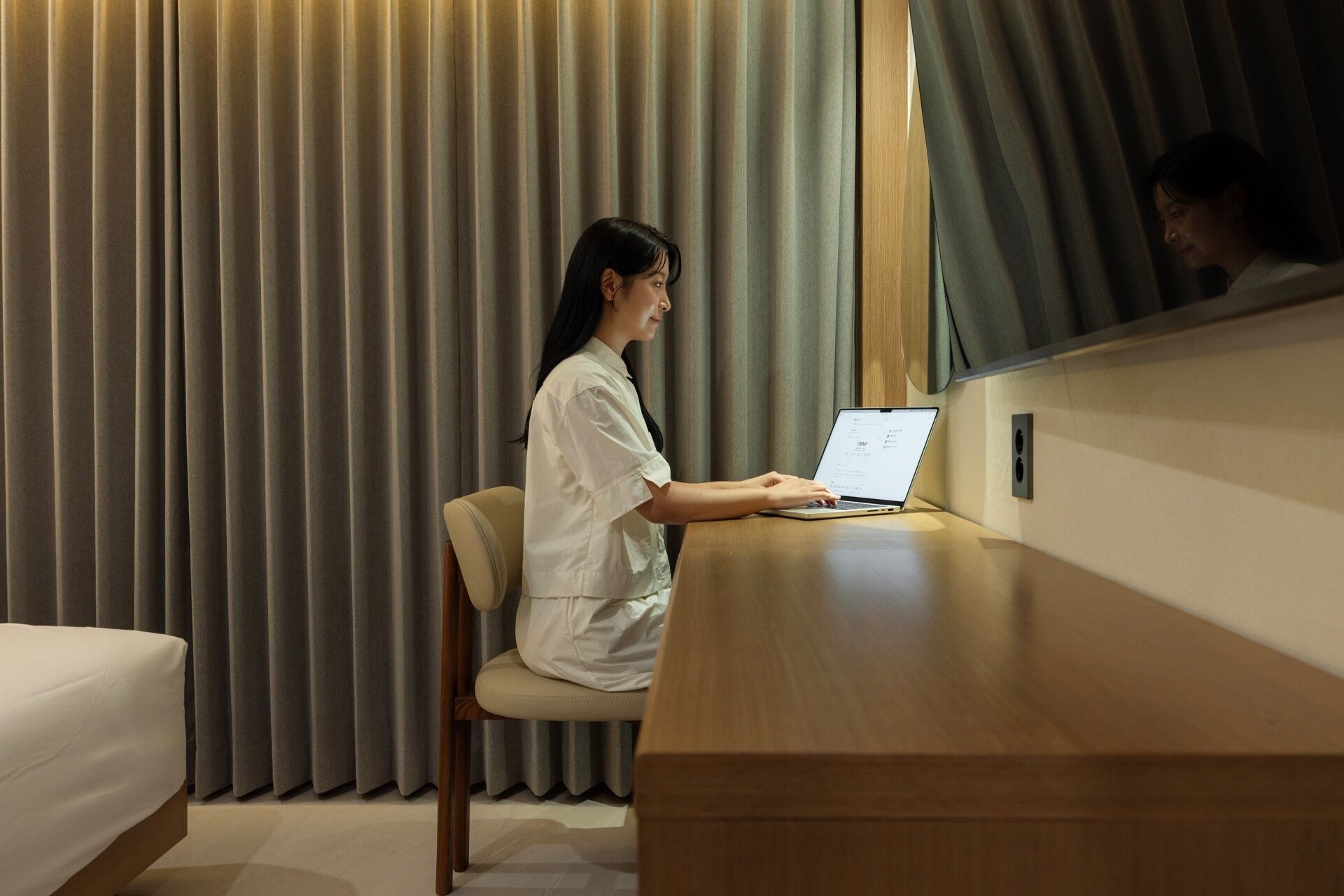 Une personne est assise à un bureau en bois, devant un ordinateur portable, dans une pièce où se trouvent un lit et des rideaux.