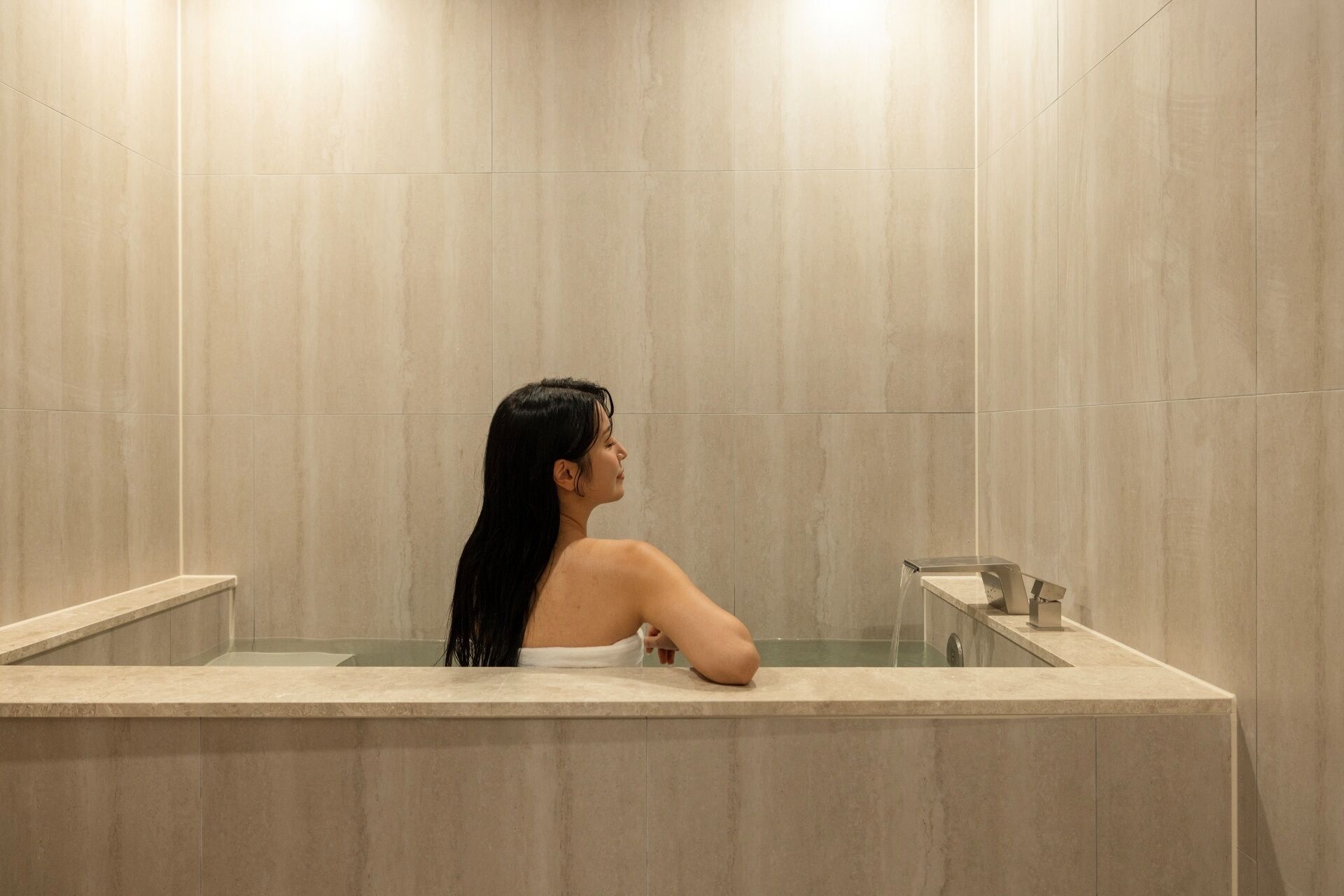 A person in a bathtub with a marble wall and a small mirror.