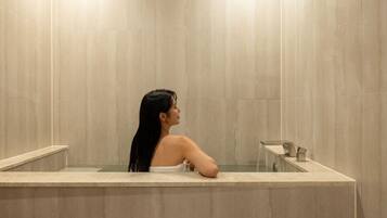A person in a bathtub with a marble wall and a small mirror.