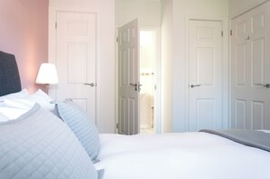 Quarto de hotel com uma cama, uma mesa de cabeceira, uma luminária e duas portas que dão para outros espaços.