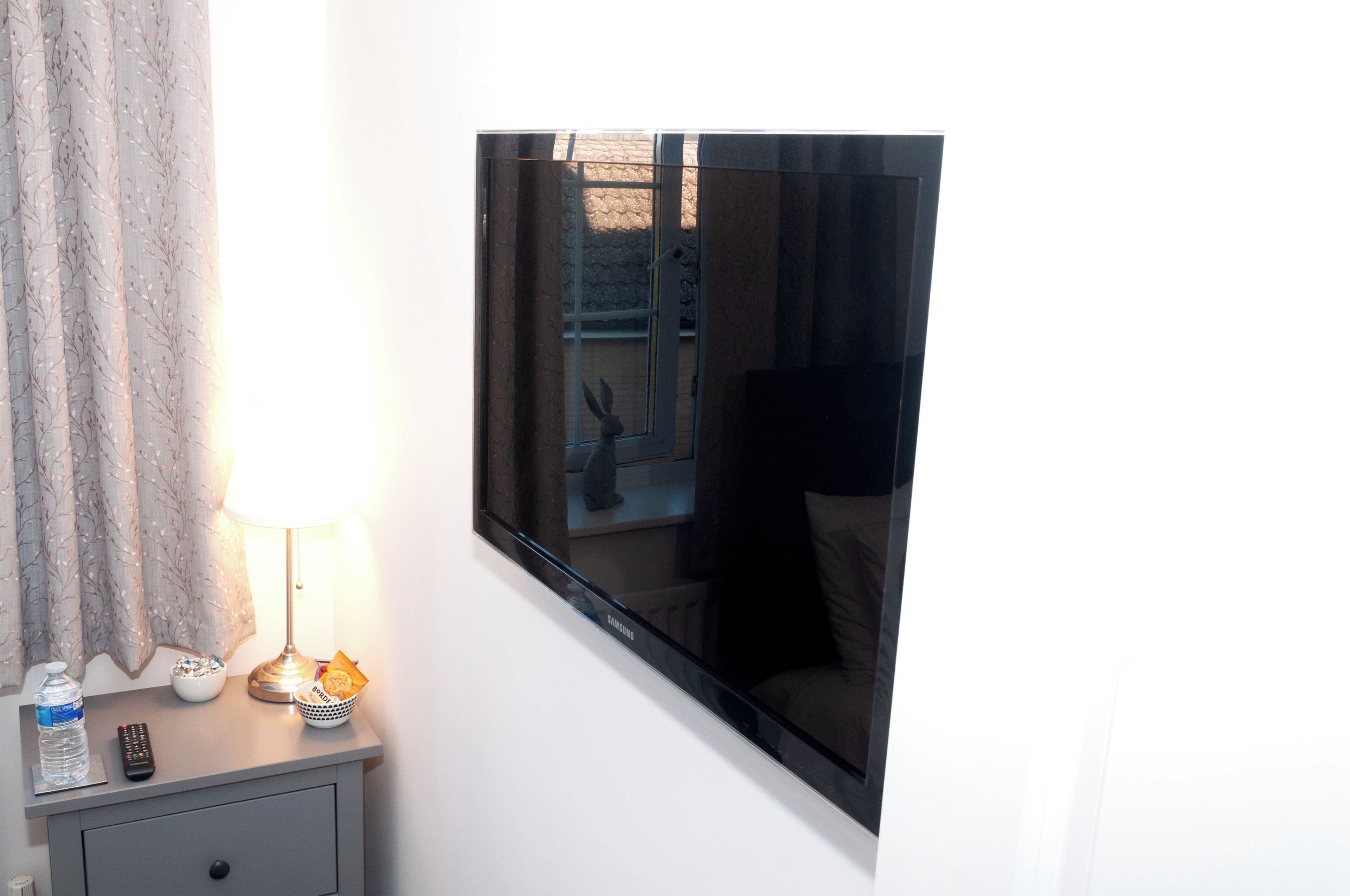 Une chambre dotée d’un grand téléviseur à écran plat fixé au mur, d’une table de chevet avec une lampe, d’une bouteille d’eau et d’une télécommande.