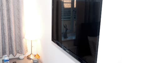 A room with a large flat-screen TV mounted on the wall, a bedside table with a lamp, a bottle of water, and a remote control.
