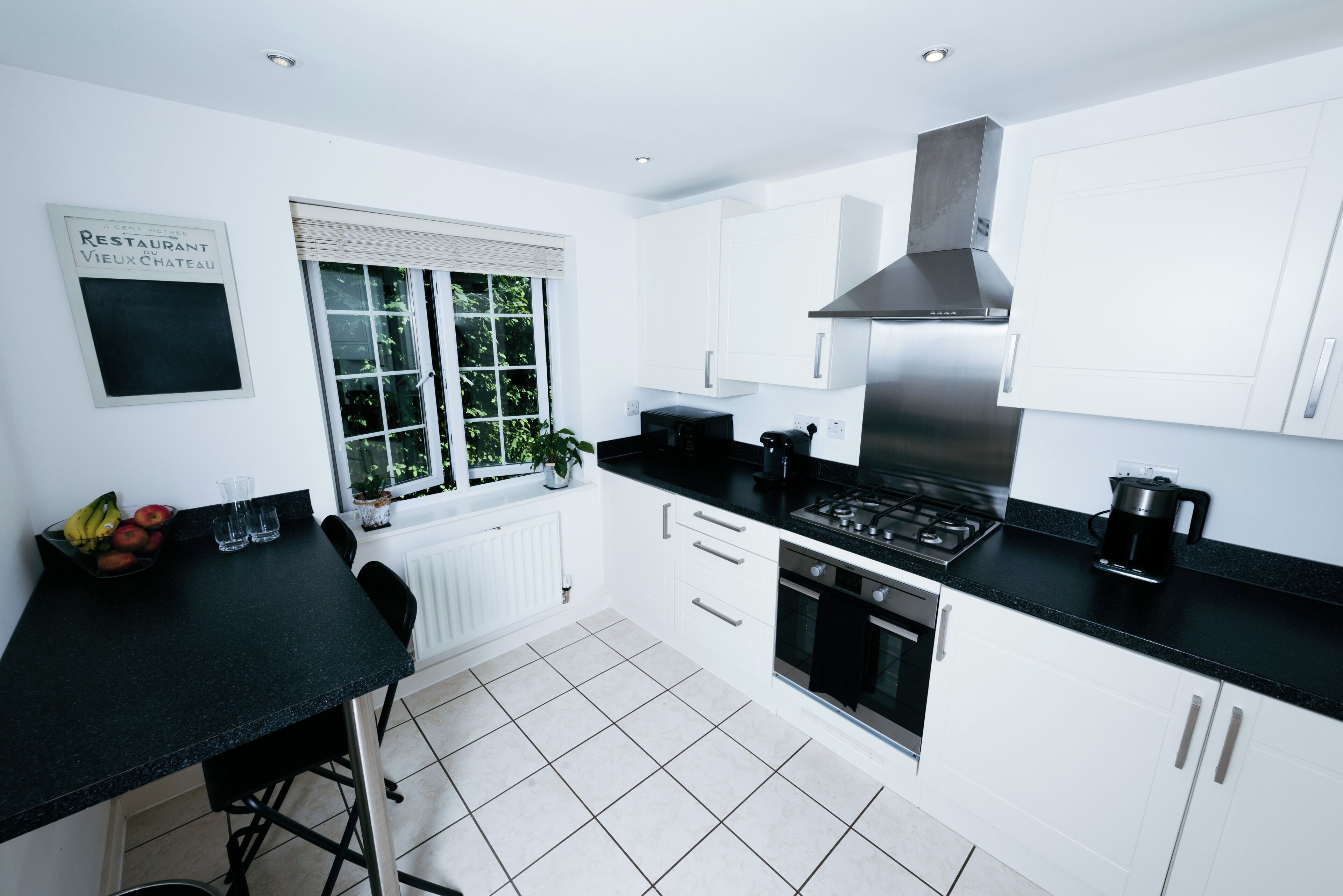 A modern kitchen with white cabinets, a black countertop, a stainless steel range hood, and a window with blinds.