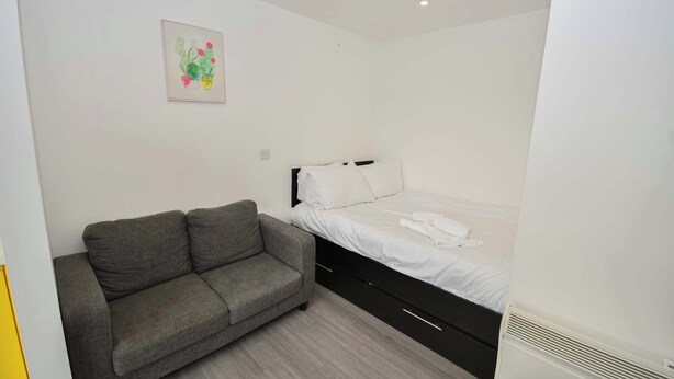 A small, clean room with a grey sofa, a single bed with white bedding, and a simple wall-mounted light fixture. - Modern & Convenient Studio in Central Sheffield (Sheffield)