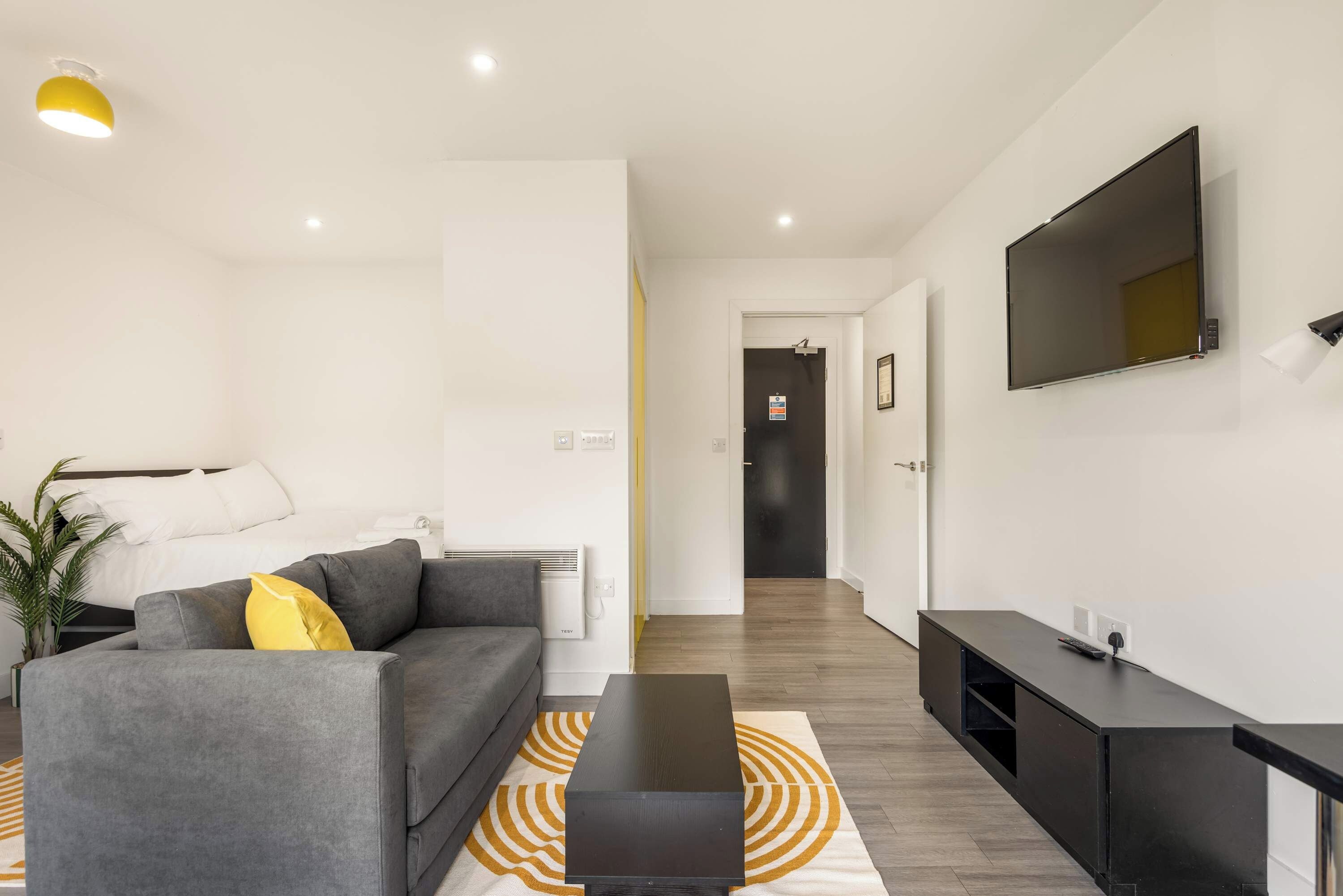 A modern living room with a grey sofa, a coffee table, a flat-screen TV mounted on the wall, and a hallway leading to a dark door.