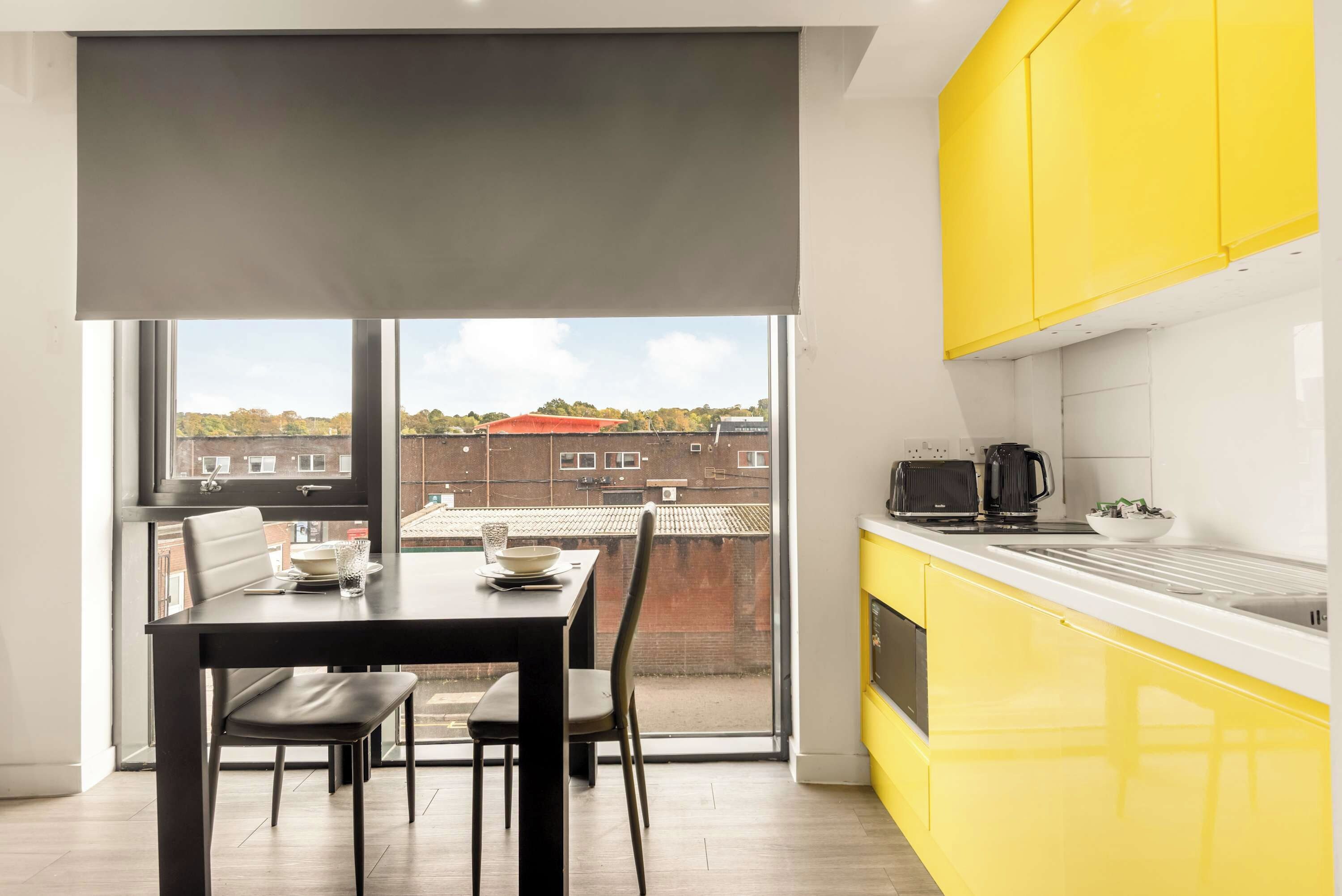 Une cuisine moderne avec des meubles de couleur jaune, un coin repas avec une table et des chaises, et une grande fenêtre offrant une vue sur les bâtiments et les arbres.