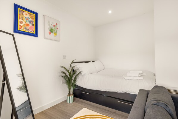 Een moderne slaapkamer met een bed, een kamerplantje, een grijze bank en twee ingelijste kunstwerken aan de muur.