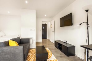 Een moderne hotelkamer met een grijze bank, een zwarte salontafel, een aan de muur bevestigde televisie en een gang naar een deur.