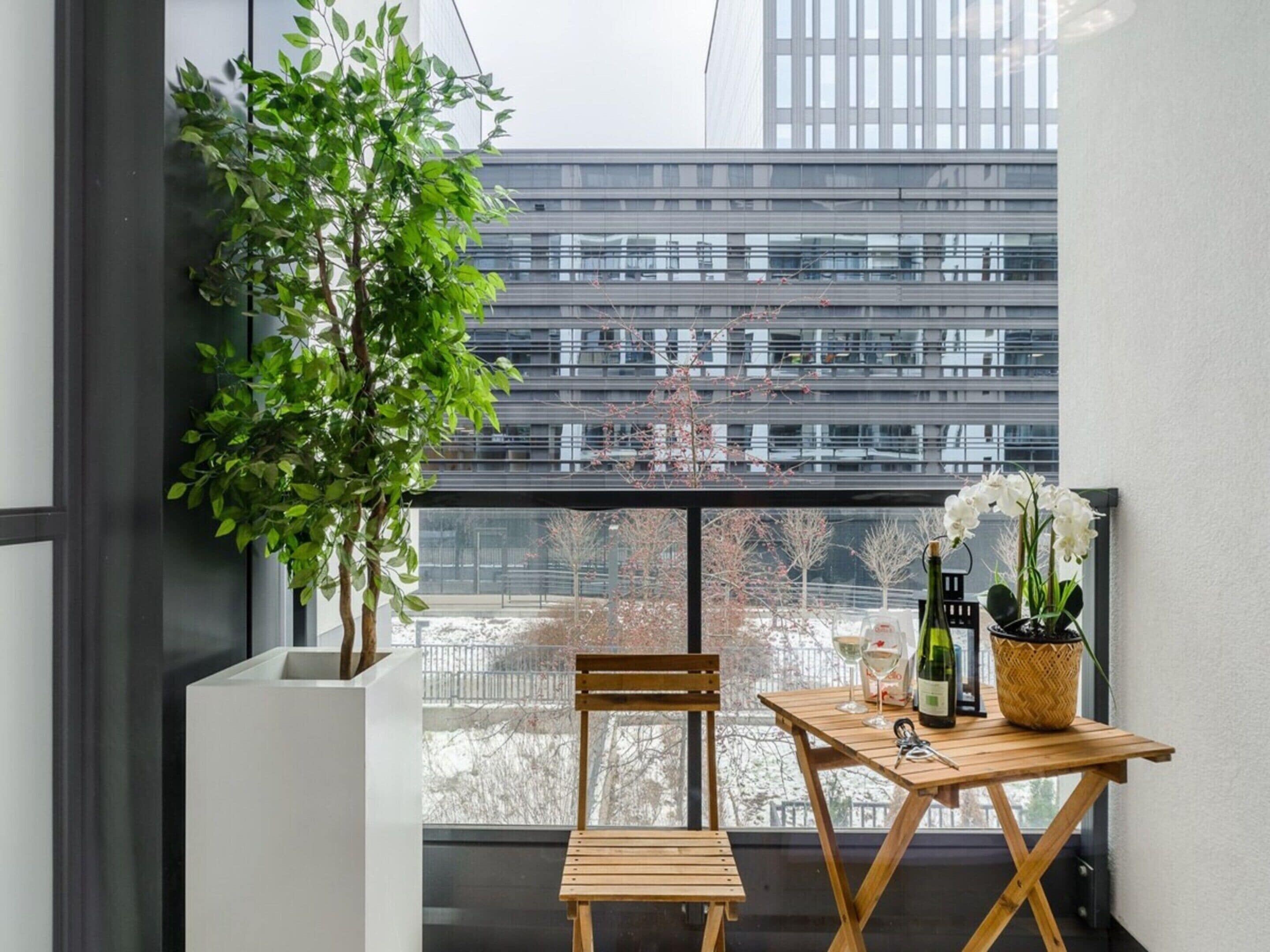 A balcony with a wooden table, a chair, a potted plant, and a view of a modern building.