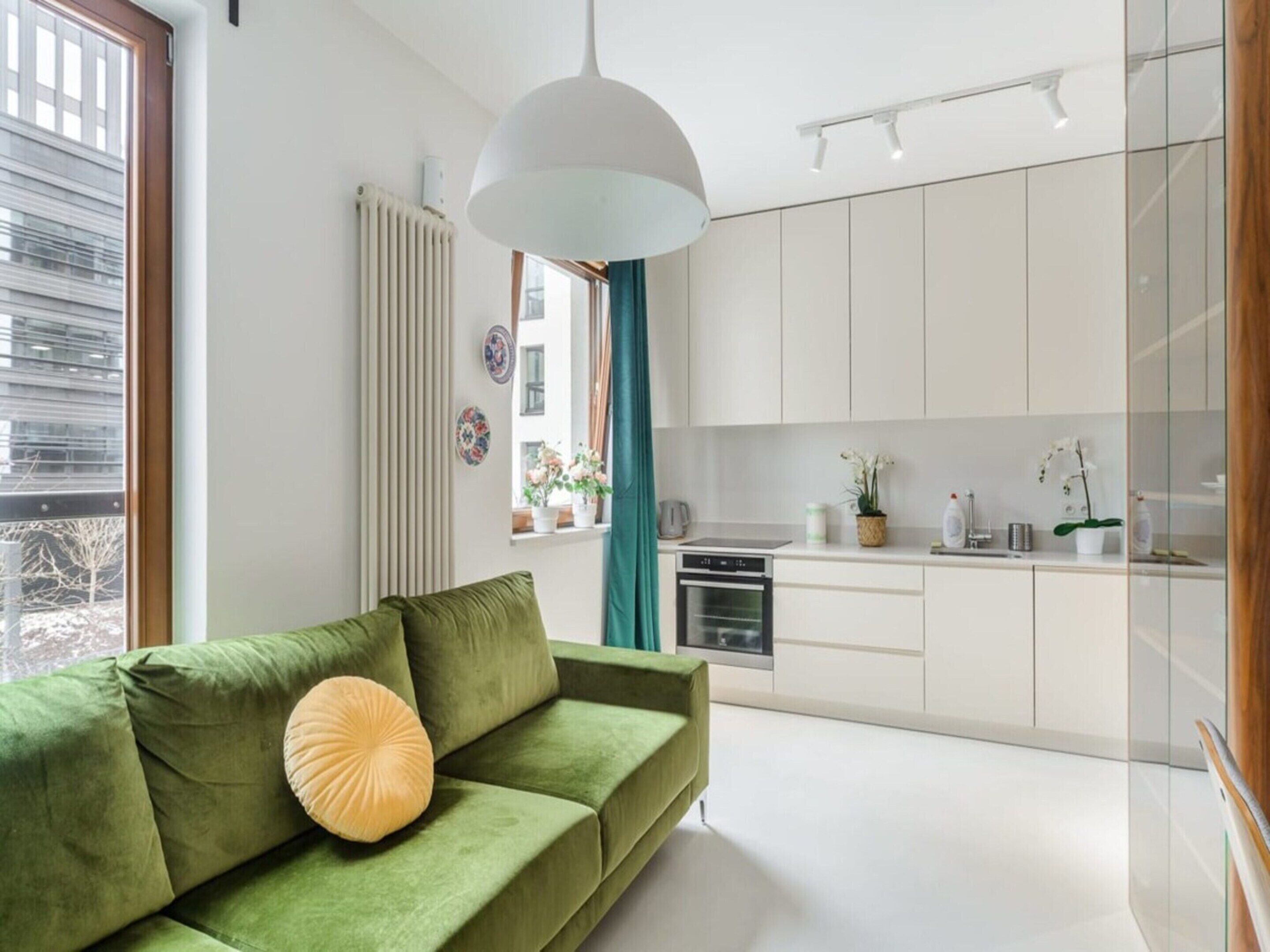 A modern living room with a green sofa, a pendant light, and a view of a kitchen with white cabinets.