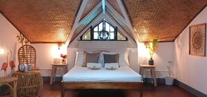 A bedroom with a bed centered under a sloped ceiling, flanked by two nightstands with lamps, a window with a wreath, and a wall-mounted artwork. - Kerusso Haven (Puerto Princesa)