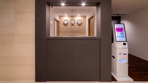 Zona de recepción con quiosco digital, pared de madera con relojes y suelo de baldosas.