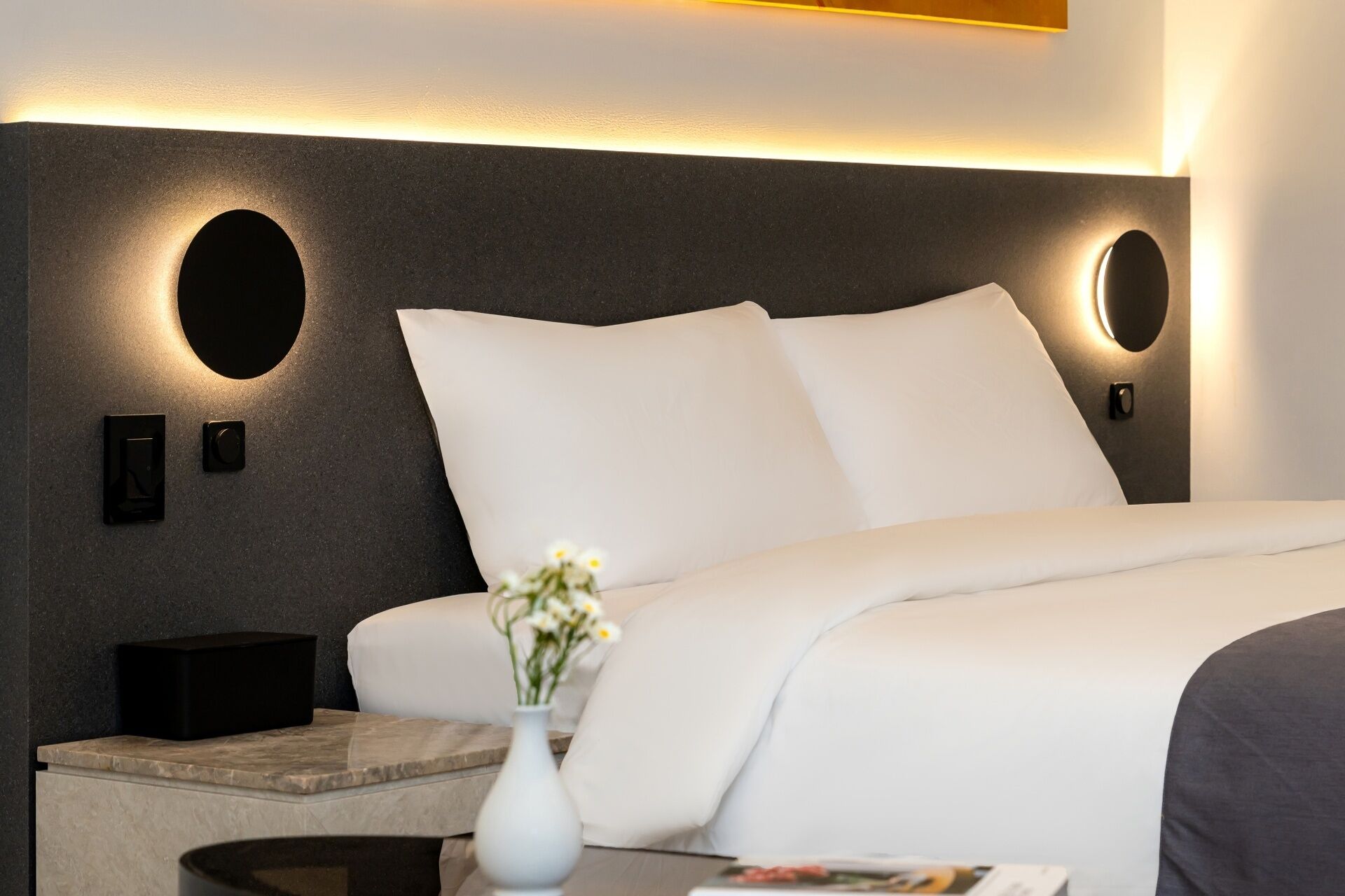 Una habitación de hotel moderna con una cama grande, una mesita de noche, un florero con flores e iluminación en la pared.