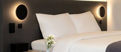 Una habitación de hotel moderna con una cama grande, una mesita de noche, un florero con flores e iluminación en la pared.