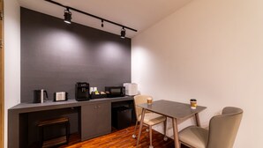 Una kitchenette compacta con cafetera, microondas y tostadora. Hay una pequeña mesa con dos sillas y una taza de café. El suelo es de madera y las paredes son lisas.
