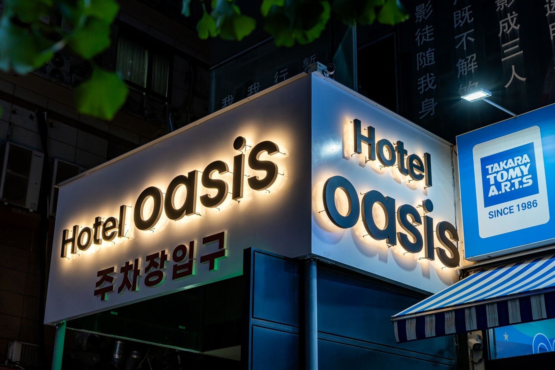 A hotel sign with illuminated letters spelling 'Hotel Oasis' and a smaller sign indicating 'Takara Tommy A.R.T.S' since 1986.