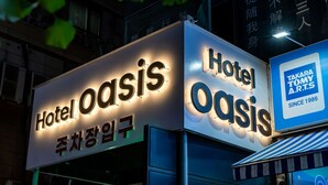Un letrero de hotel con letras iluminadas que dicen 'Hotel Oasis' y un letrero más pequeño que indica 'Takara Tommy A.R.T.S' desde 1986.