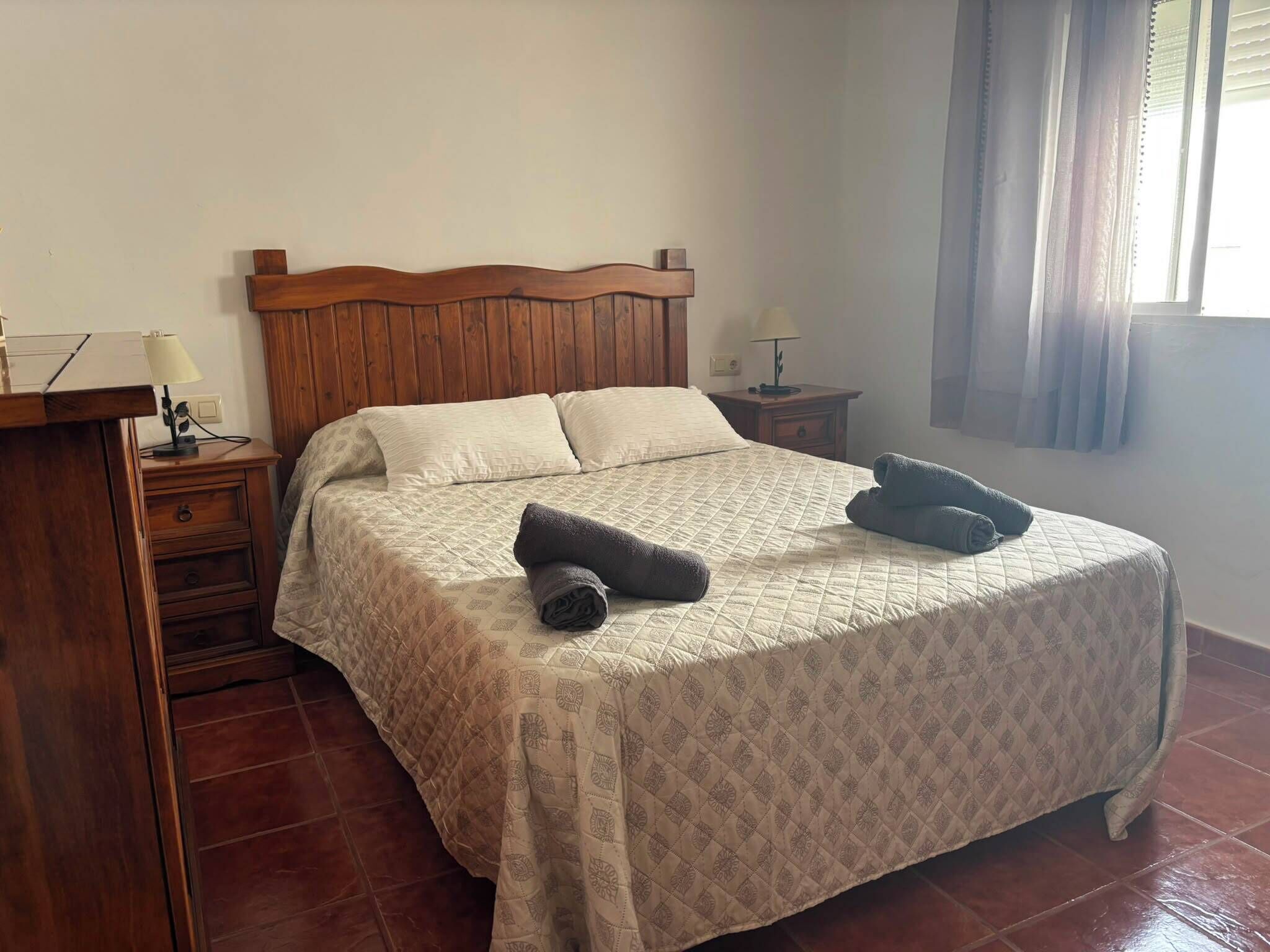 A bedroom with a wooden headboard, a bed with a quilted bedspread, two pillows, a nightstand with a lamp, and a window with curtains.