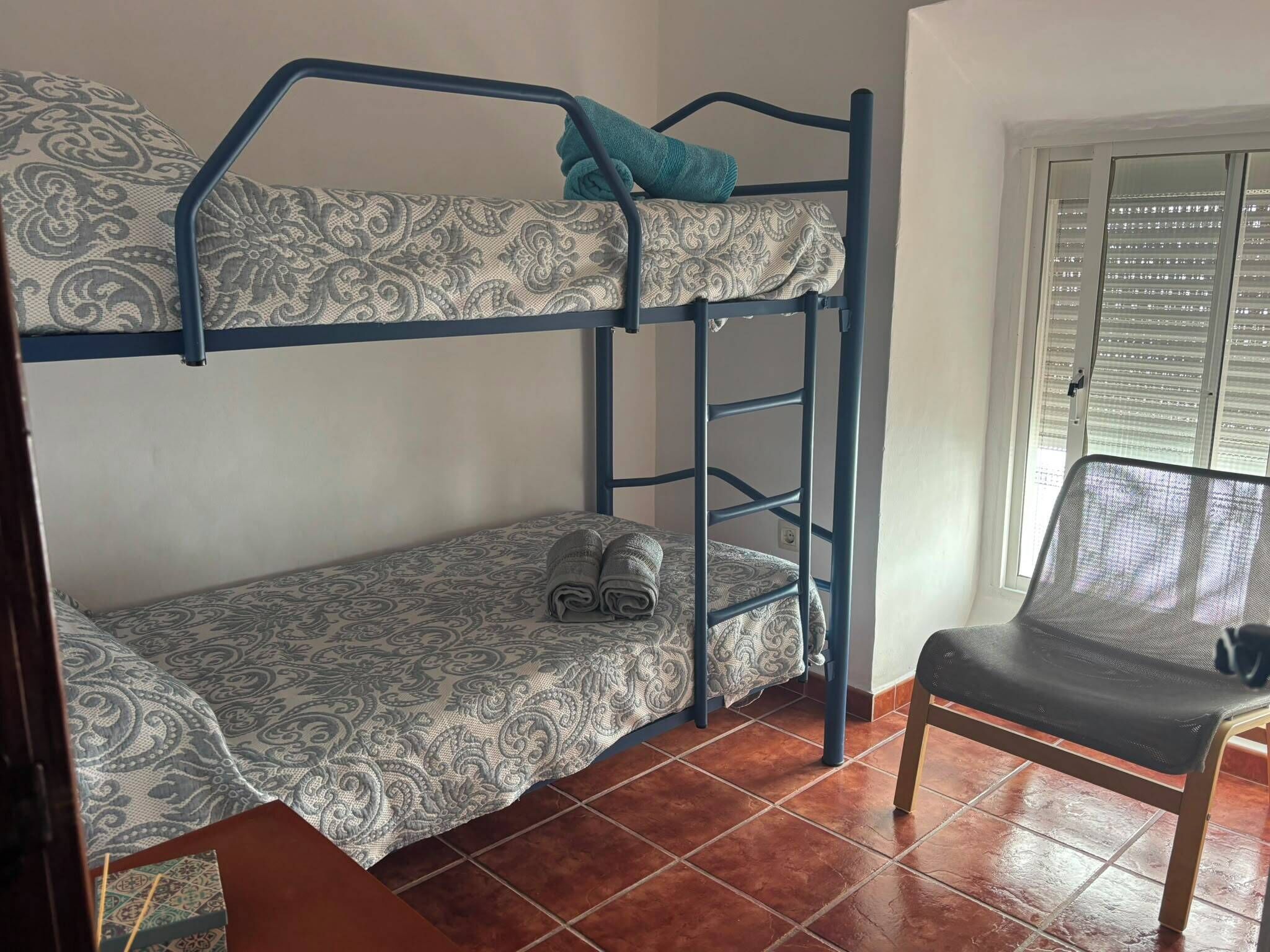 A bunk bed with patterned bedding, a chair with a mesh backrest, and a window with blinds.