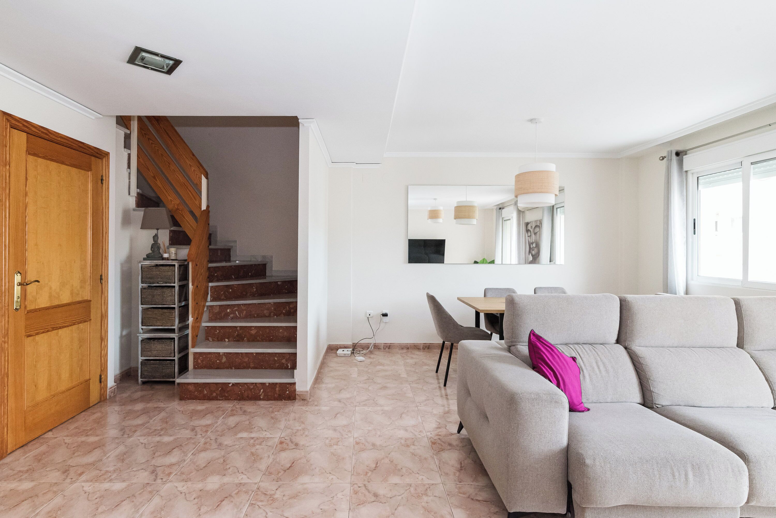 A modern living room with a grey sofa, a wooden door, a staircase, and a dining area with a table and chairs.