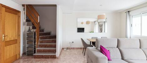 Una sala de estar moderna con un sofá gris, una puerta de madera, una escalera y un comedor con mesa y sillas.
