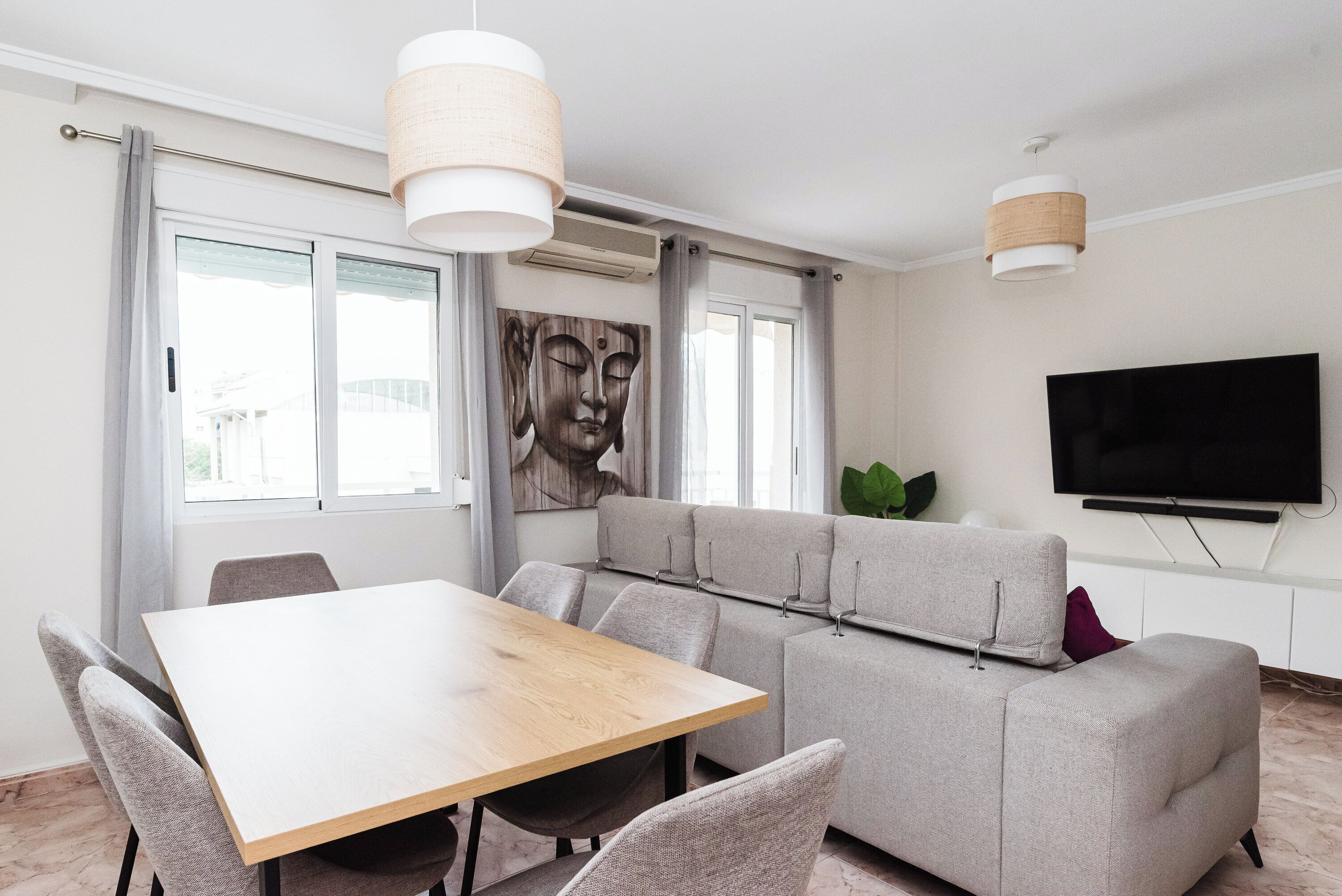 A modern living room with a wooden dining table, grey sofa, and a flat-screen TV mounted on the wall.
