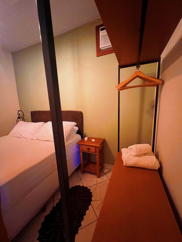 A hotel room with a bed, a bedside table, a hanging coat rack, and a mirror on the wall.
