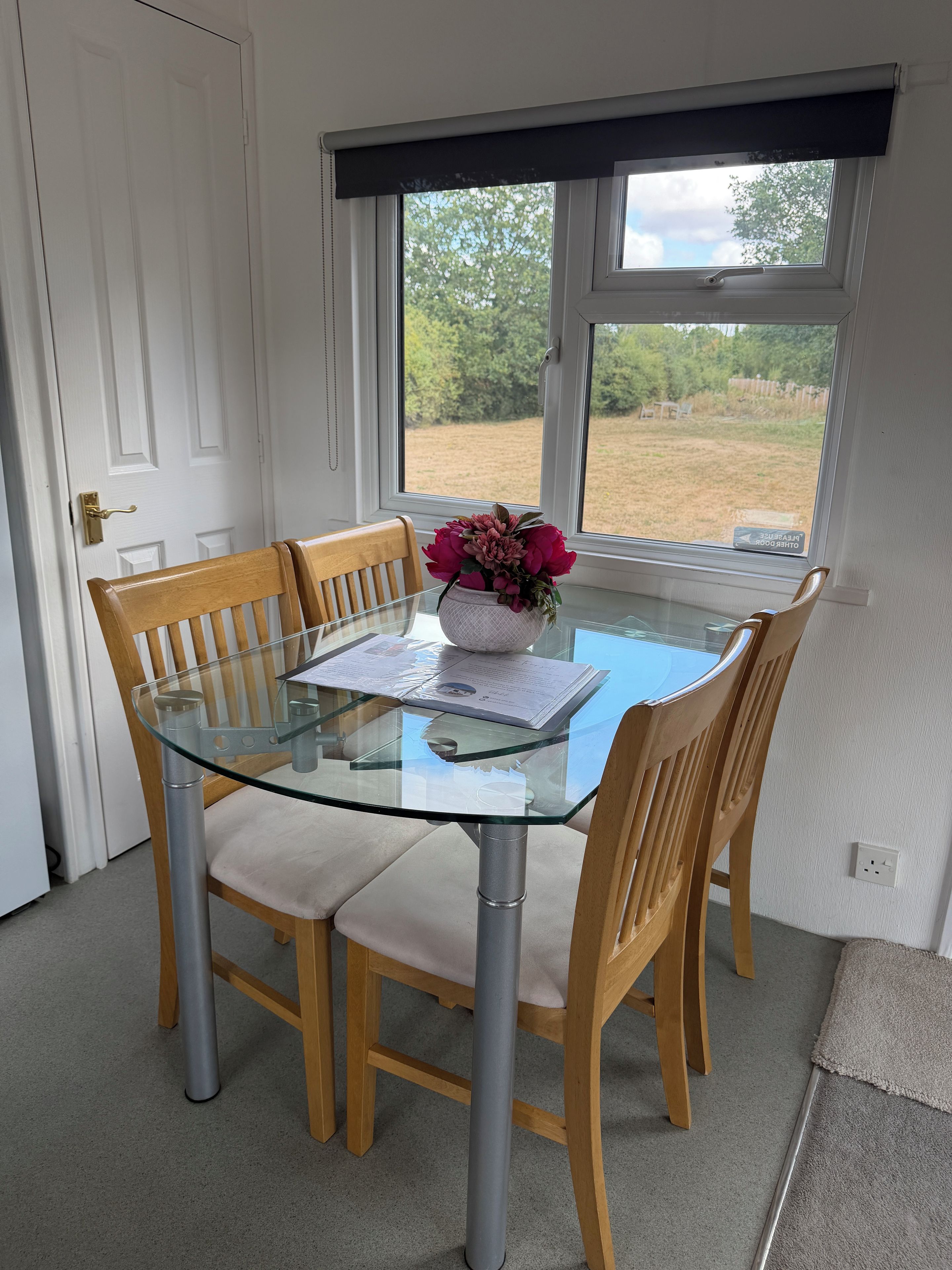 A dining area with a glass table surrounded by wooden chairs, a vase with flowers, and a view of the outdoors through a window.