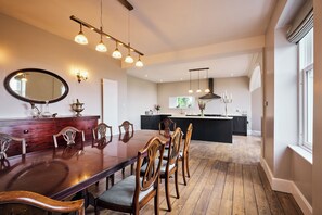 Uma sala de jantar com mesa e cadeiras de madeira, uma cozinha ao fundo e luminárias pendentes.