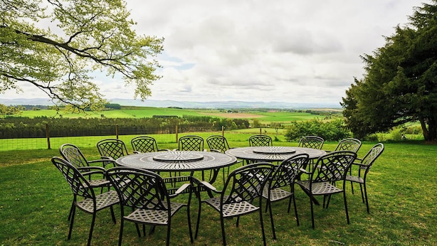 Un espace extérieur circulaire aménagé avec des chaises et des tables en métal, entouré d’un champ verdoyant offrant une vue sur un paysage lointain.