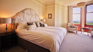 Un grand lit avec une tête de lit décorée, une table de chevet avec une lampe, une chaise, une table et une vue sur l’extérieur par une fenêtre.