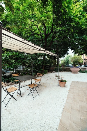 Uma área externa com mesas e cadeiras sob uma pérgola sombreada, cercada por vegetação e um caminho de cascalho.