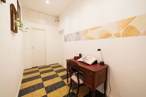 Um corredor com piso xadrez, uma escrivaninha de madeira com uma luminária e uma porta.