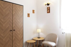 Quarto com guarda-roupa em padrão losango, uma pequena mesa redonda com abajur, uma cadeira e dois quadros pendurados na parede.